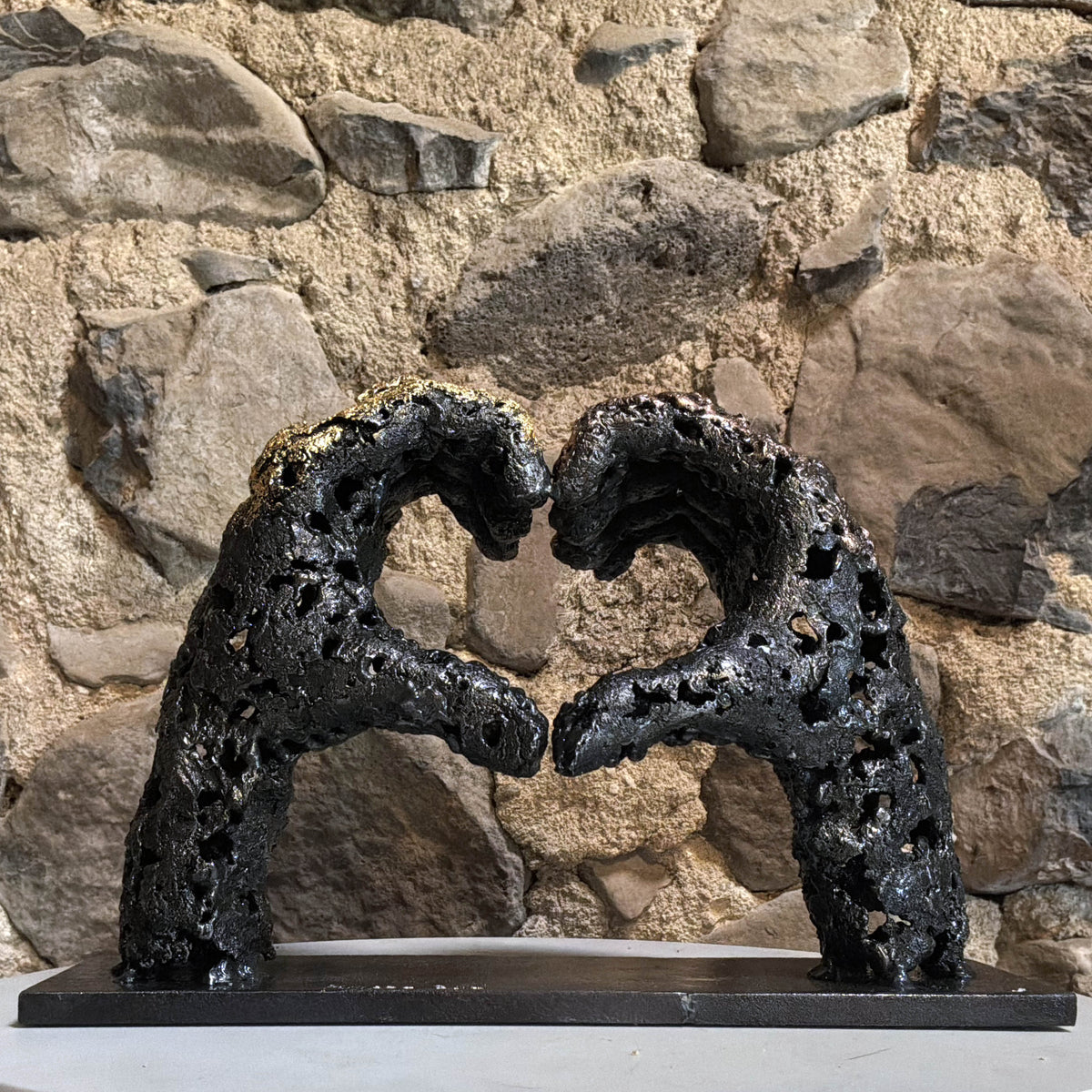 Sculpture « Un échange », deux mains formant un cœur en acier et bronze, présentée dans un intérieur au mur de pierre