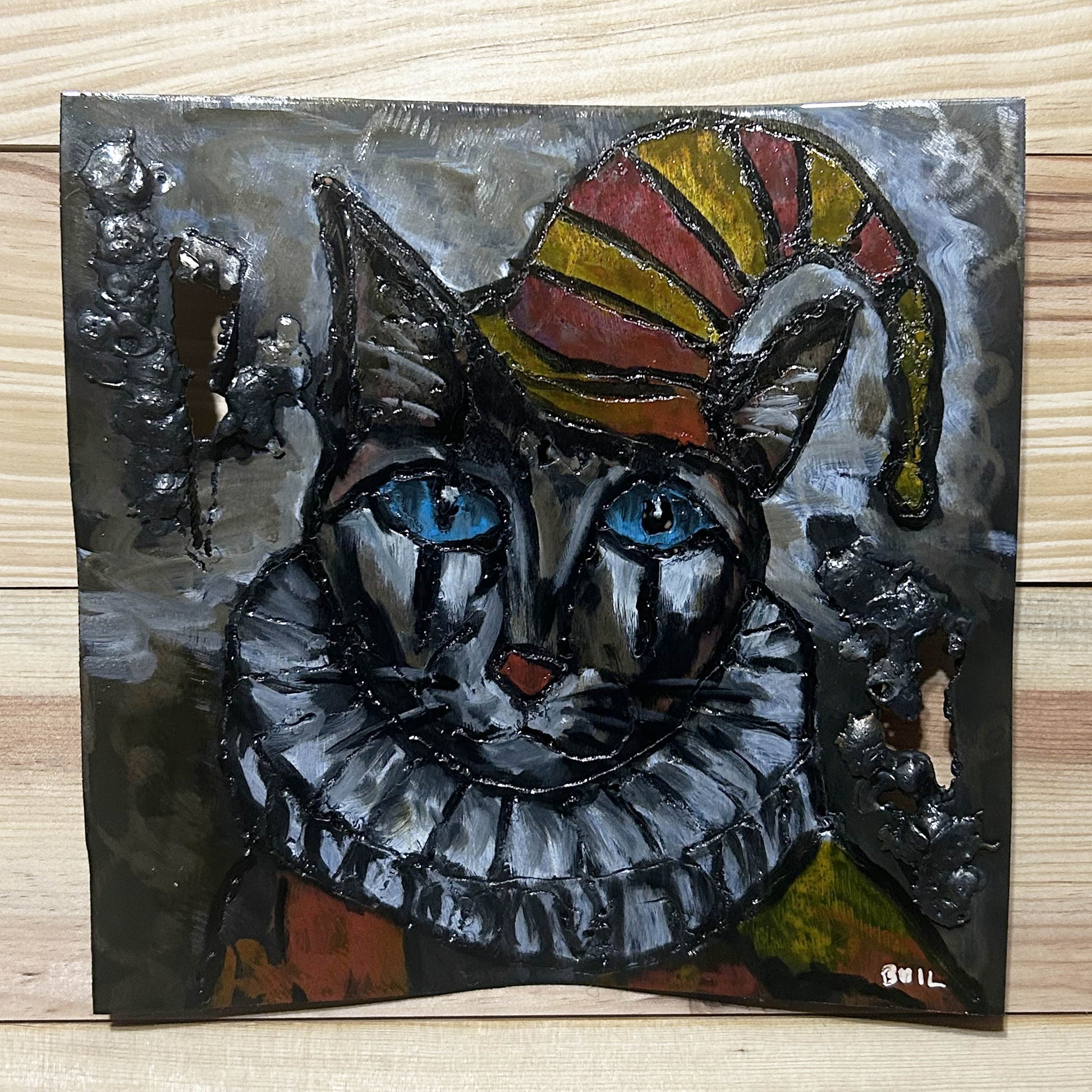 Sculpture murale carrée de 25x25 cm représentant un chat arlequin aux yeux bleus intenses, portant une collerette blanche et un chapeau rayé rouge et jaune. L’œuvre est réalisée en acier texturé et pigments, avec un fond gris brumeux et métallisé