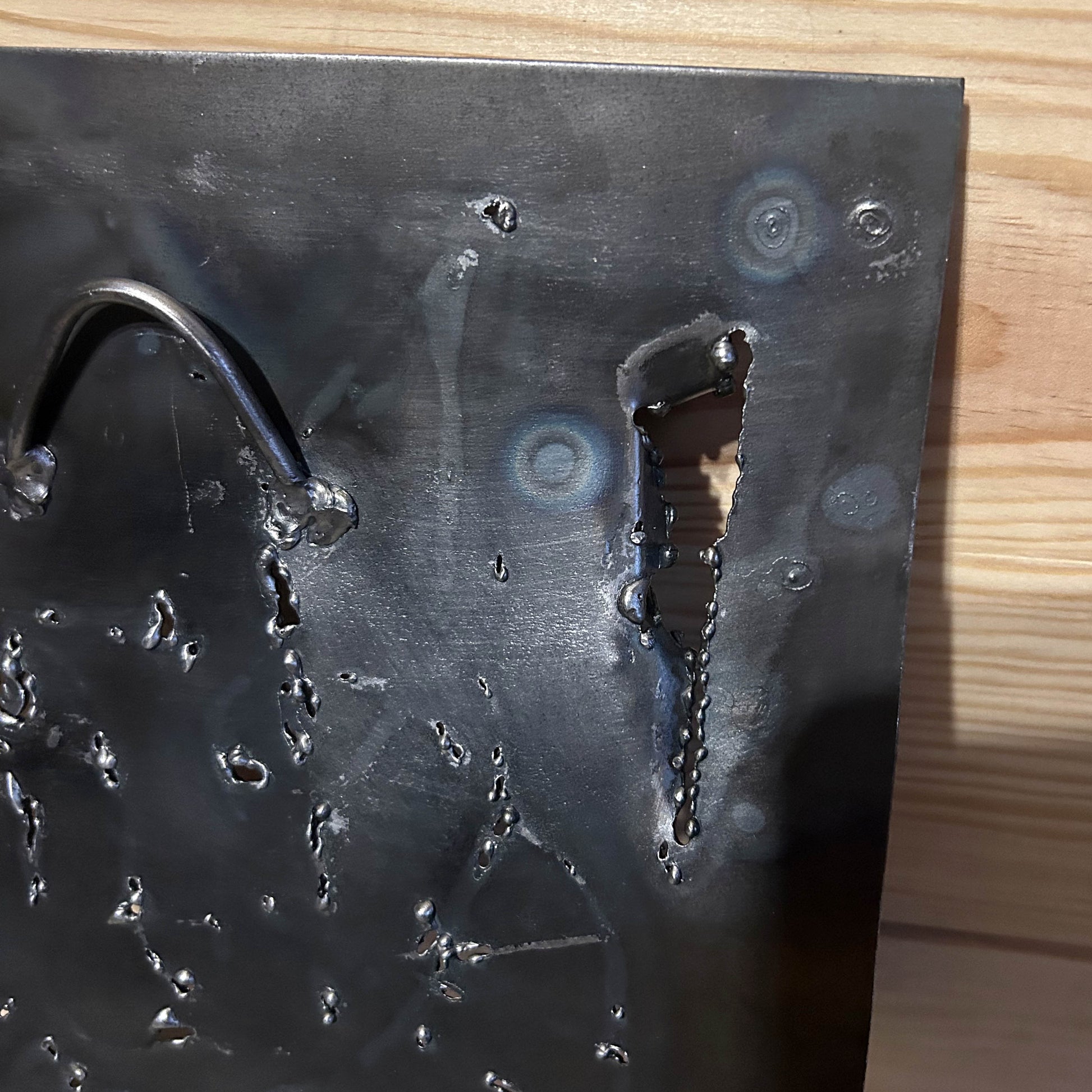 Sculpture murale carrée de 25x25 cm représentant un chat arlequin aux yeux bleus intenses, portant une collerette blanche et un chapeau rayé rouge et jaune. L’œuvre est réalisée en acier texturé et pigments, avec un fond gris brumeux et métallisé