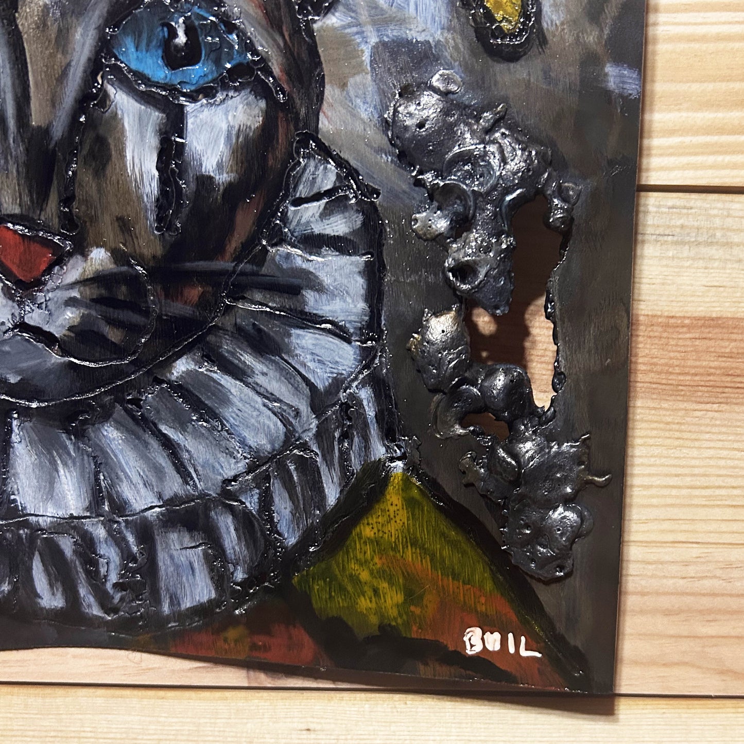 Sculpture murale carrée de 25x25 cm représentant un chat arlequin aux yeux bleus intenses, portant une collerette blanche et un chapeau rayé rouge et jaune. L’œuvre est réalisée en acier texturé et pigments, avec un fond gris brumeux et métallisé