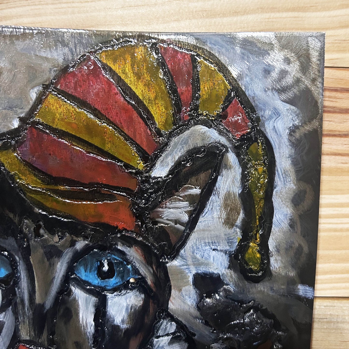 Sculpture murale carrée de 25x25 cm représentant un chat arlequin aux yeux bleus intenses, portant une collerette blanche et un chapeau rayé rouge et jaune. L’œuvre est réalisée en acier texturé et pigments, avec un fond gris brumeux et métallisé