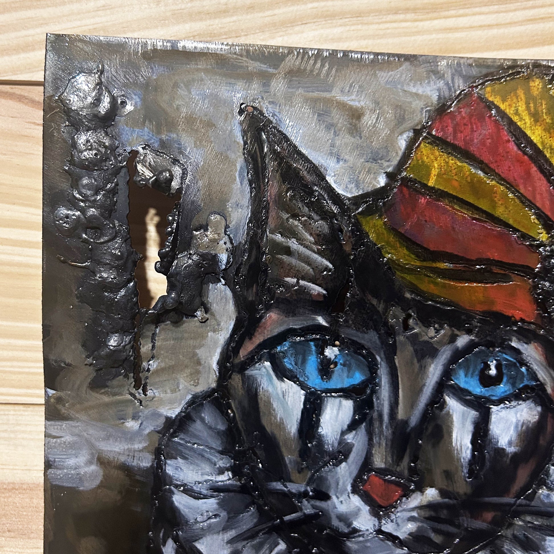 Sculpture murale carrée de 25x25 cm représentant un chat arlequin aux yeux bleus intenses, portant une collerette blanche et un chapeau rayé rouge et jaune. L’œuvre est réalisée en acier texturé et pigments, avec un fond gris brumeux et métallisé