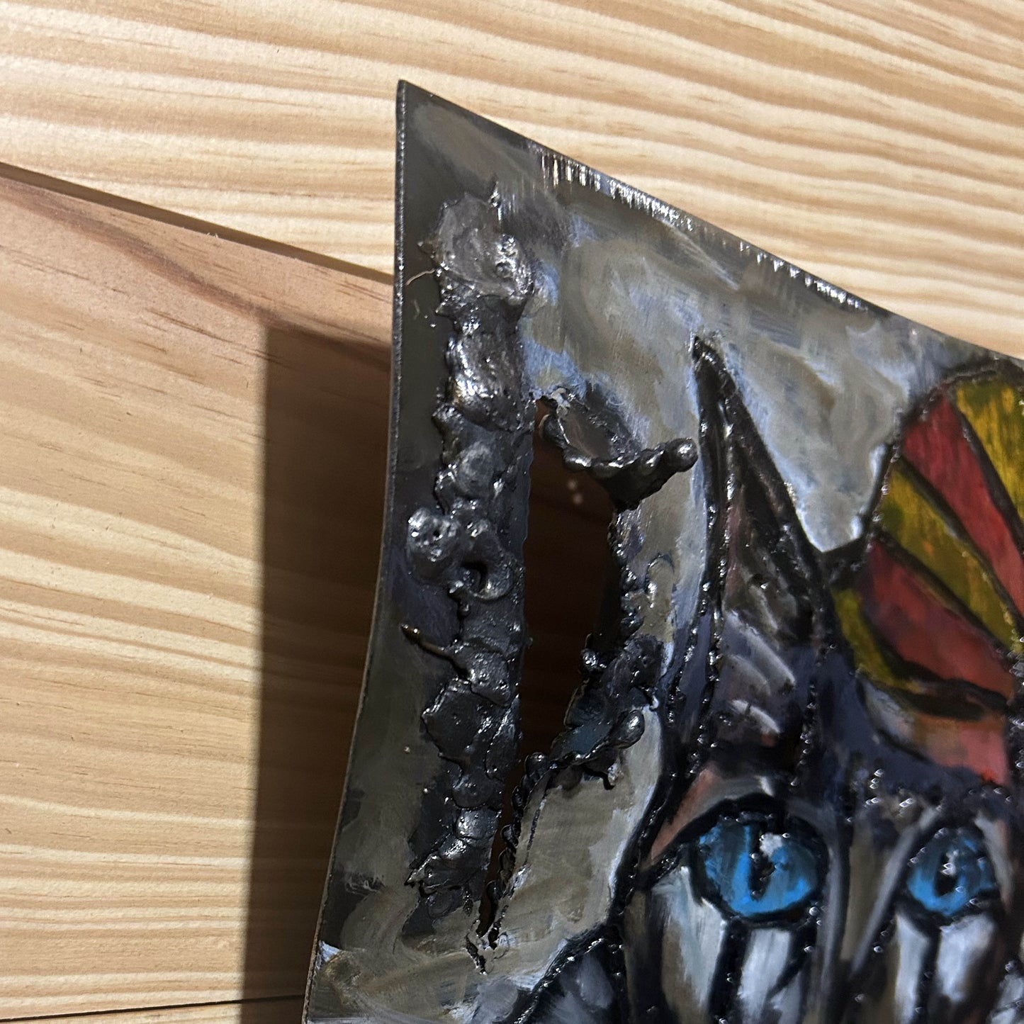 Sculpture murale carrée de 25x25 cm représentant un chat arlequin aux yeux bleus intenses, portant une collerette blanche et un chapeau rayé rouge et jaune. L’œuvre est réalisée en acier texturé et pigments, avec un fond gris brumeux et métallisé