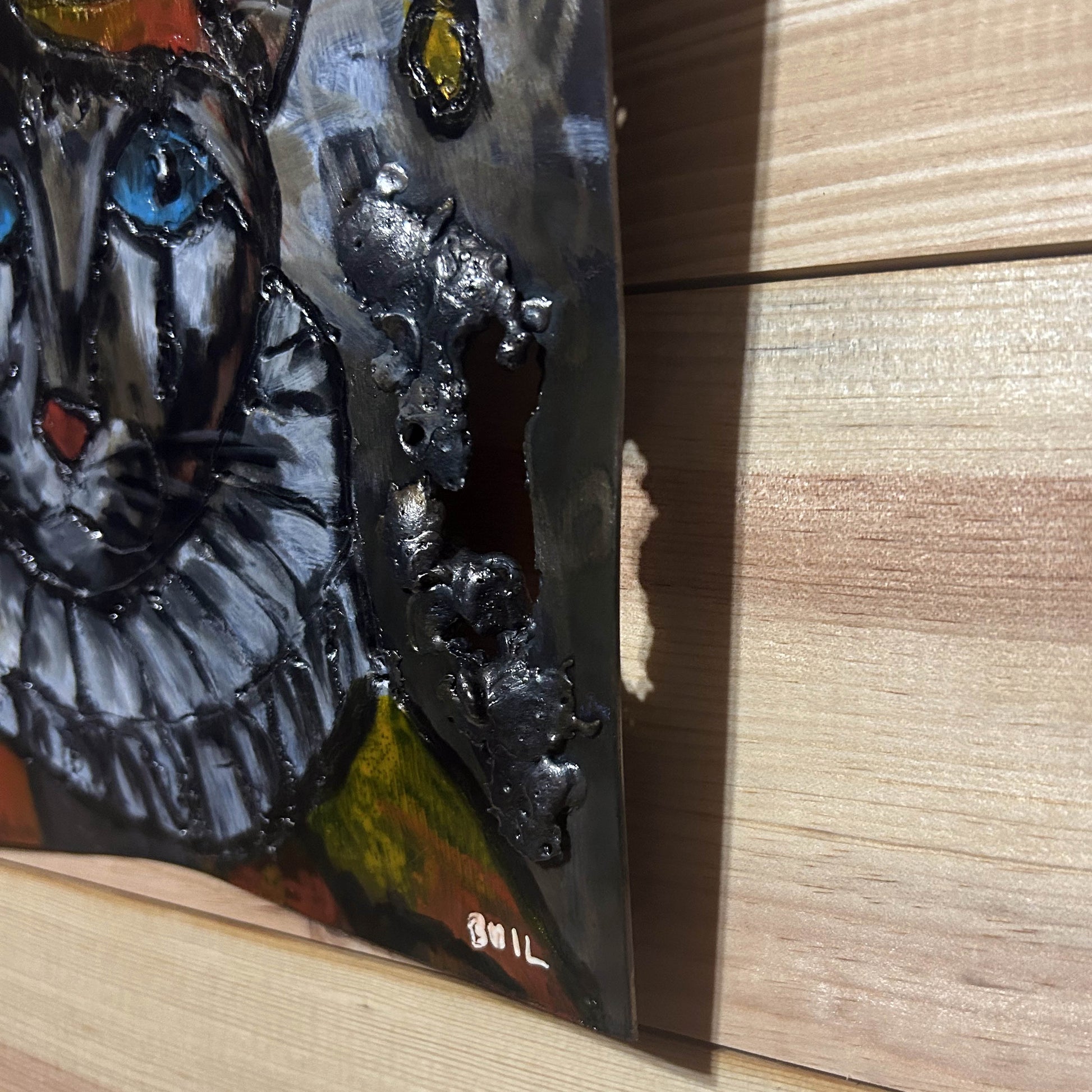 Sculpture murale carrée de 25x25 cm représentant un chat arlequin aux yeux bleus intenses, portant une collerette blanche et un chapeau rayé rouge et jaune. L’œuvre est réalisée en acier texturé et pigments, avec un fond gris brumeux et métallisé