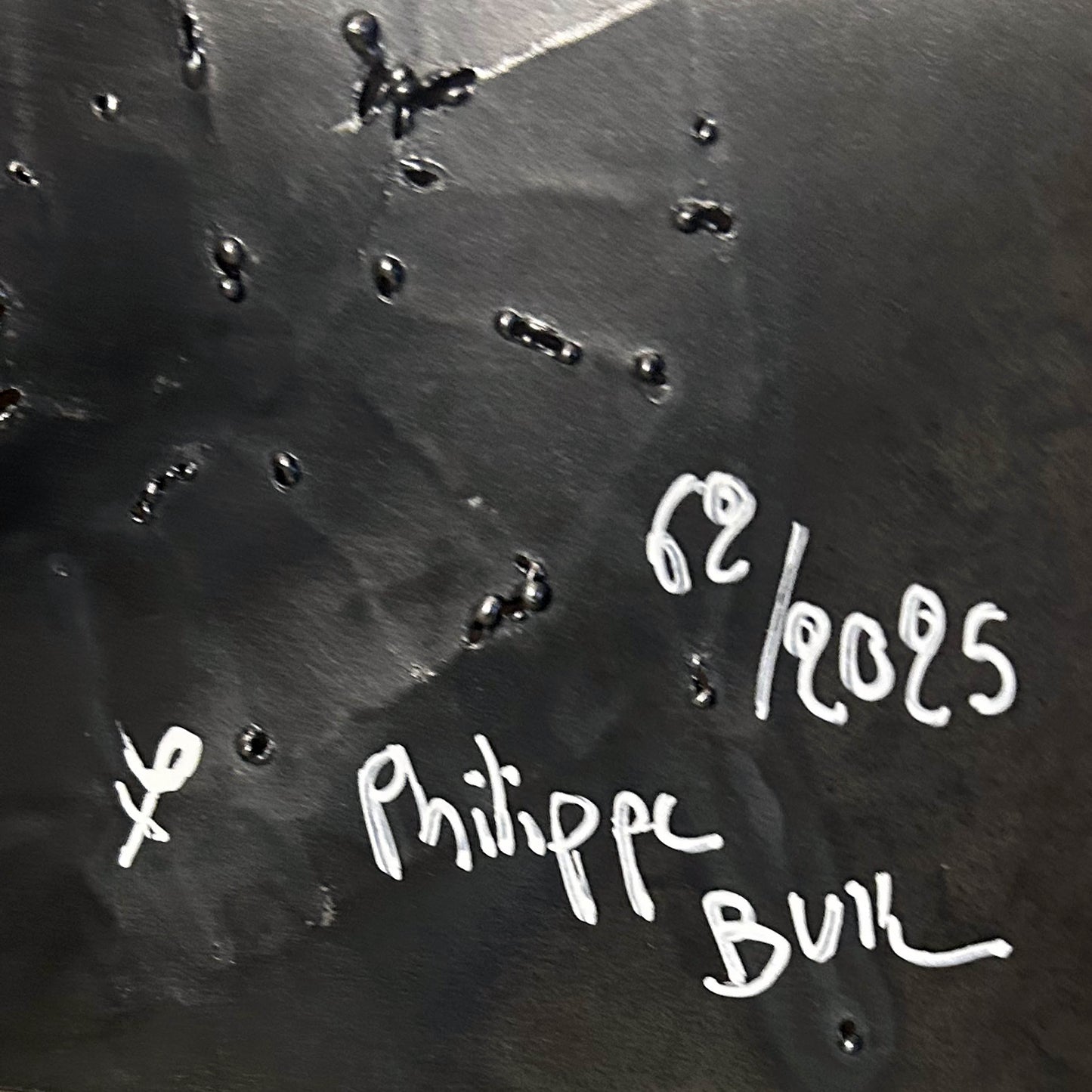 Sculpture murale carrée de 25x25 cm représentant un chat arlequin aux yeux bleus intenses, portant une collerette blanche et un chapeau rayé rouge et jaune. L’œuvre est réalisée en acier texturé et pigments, avec un fond gris brumeux et métallisé