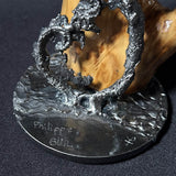 Sculpture contemporaine représentant une salamandre en dentelle de métal (acier), fixée sur un socle métallique circulaire et appuyée contre un morceau de bois naturel, œuvre de Philippe Buil, sur fond sombre.