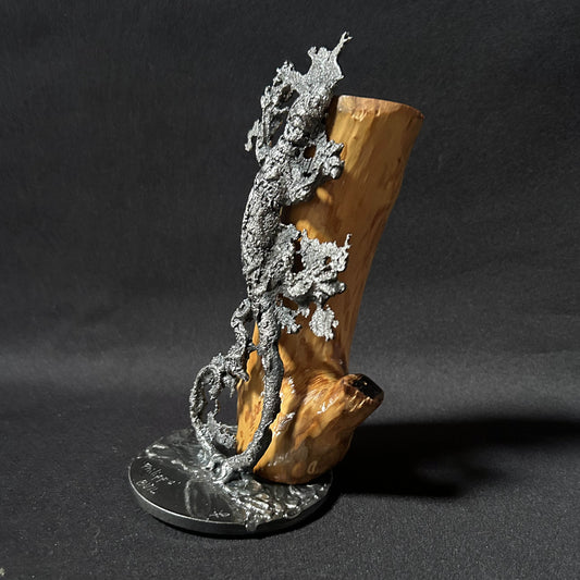 Sculpture contemporaine représentant une salamandre en dentelle de métal (acier), fixée sur un socle métallique circulaire et appuyée contre un morceau de bois naturel, œuvre de Philippe Buil, sur fond sombre.