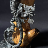 Sculpture contemporaine représentant une salamandre en dentelle de métal (acier), fixée sur un socle métallique circulaire et appuyée contre un morceau de bois naturel, œuvre de Philippe Buil, sur fond sombre.